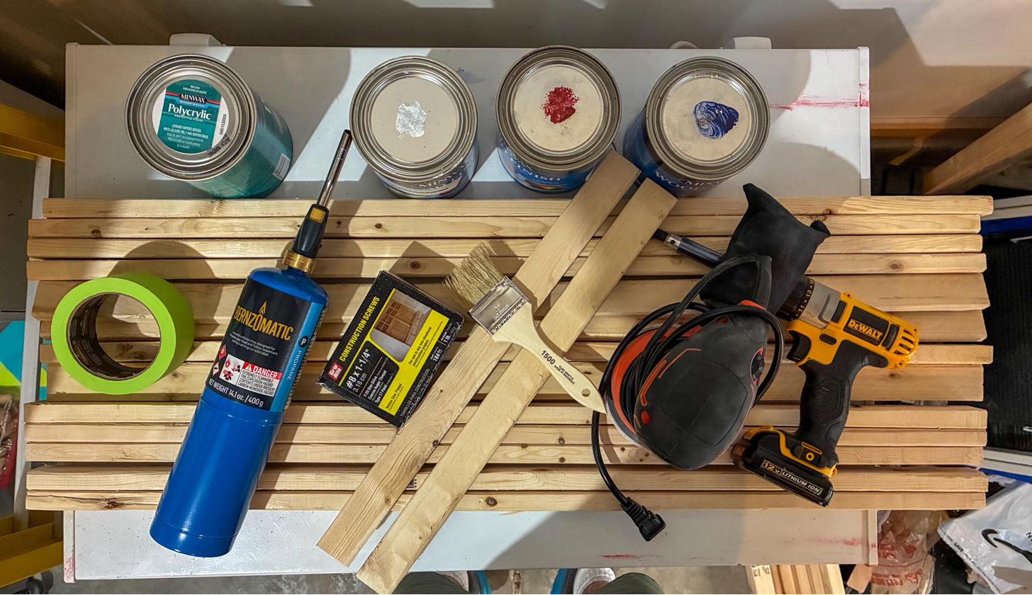 Flat-lay of the tools and materials to build a DIY wood American flag — raw pine slats, wood stain, spray paint, a star stencil, torch, clamps and a drill